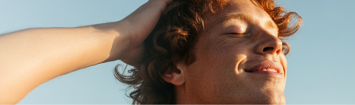 A man running his hand through his hair whilst looking up at the sun. There is only blue sky in the background